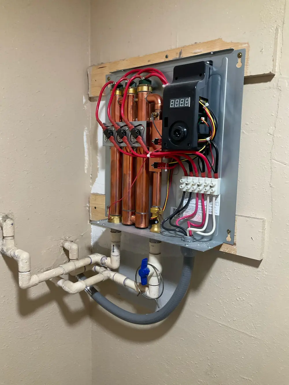 Interior view of a wall-mounted tankless water heater showing exposed copper pipes, electrical wiring, and connected PVC plumbing.