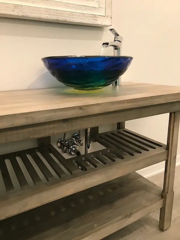 Blue glass vessel sink with waterfall faucet on a wood vanity, showing under-sink plumbing in a residential bathroom in Avila, Tampa, FL.