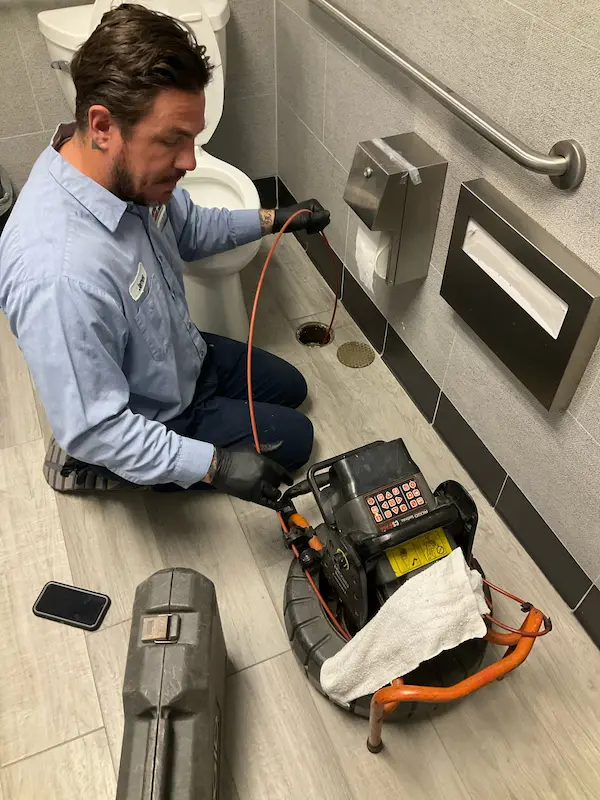 Technician clearing a clogged floor drain in a commercial restroom with a cable machine and inspection setup in Cheval, FL.