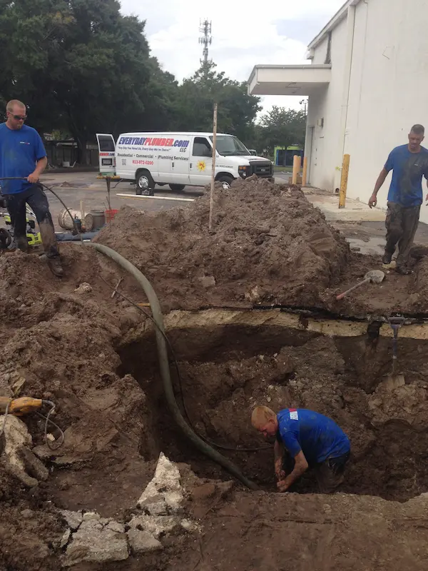 Crew excavating a large trench in a commercial parking lot to replace a broken sewer main, dewatering hose and service van on site.