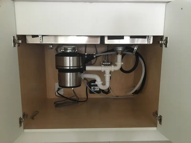 Garbage disposal installed under a double-bowl kitchen sink with PVC P-trap and dishwasher drain connection.