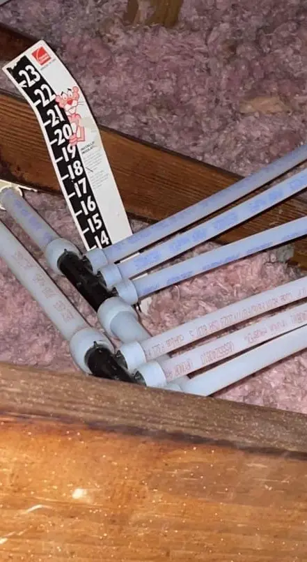 PEX repipe installation in an attic, showing multiple white flexible pipes connected with fittings and secured above pink insulation, with a temperature gauge strip nearby.