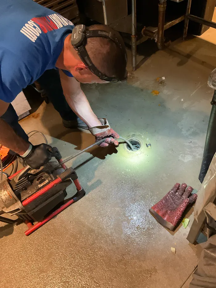 Plumber using a motorized drain auger to clear a floor drain, wearing gloves and headlamp for visibility, with coiled cable feeding into the pipe opening.