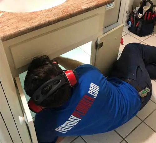 Plumber from EVERYDAYPLUMBER.com wearing protective earmuffs while repairing a leak under a kitchen sink.