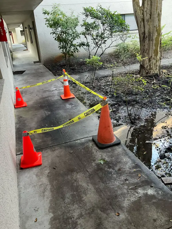 Plumbing job site with safety cones and caution tape marking a repaired section of concrete sidewalk, ensuring the area is well-marked and leveled correctly near a landscaped area.