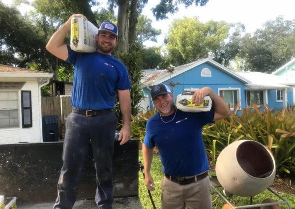 Two smiling plumbers in blue shirts carry bags of concrete mix at a Tampa job site, standing in front of residential homes and a cement mixer