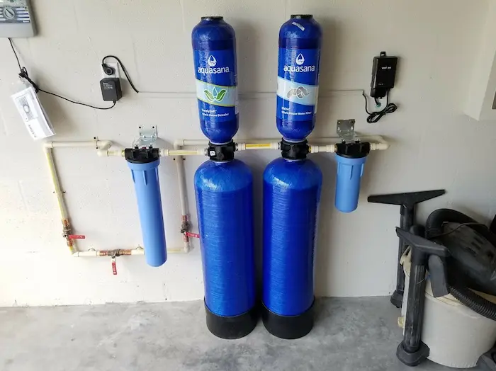 Aquasana whole-house water filtration and conditioner system with prefilters and bypass valves mounted on a garage wall.