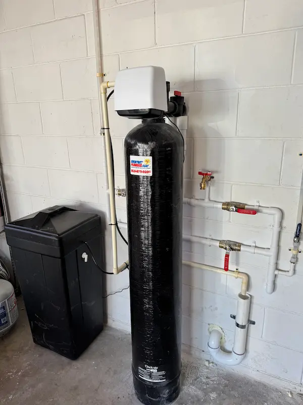 Newly installed whole-house water filtration system with a tall black tank and connected piping, set up in a garage against a white cinder block wall.