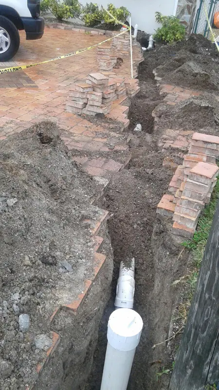 Plumbing trench in Ybor City showing replacement of an old cast iron sewer line with modern PVC piping beneath a brick driveway, with stacked bricks and caution tape marking the work area.